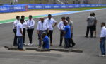 Racing team staff and engineers inspect a damaged asphalt section on the track, with two workers digging at a crack in front of them.