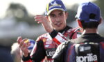 MotoGP rider in a red racing suit with a Michelin cap gestures while talking to a teammate in the pit area.