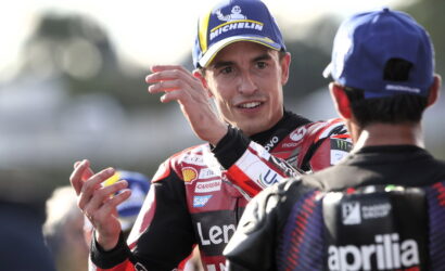 MotoGP rider in a red racing suit with a Michelin cap gestures while talking to a teammate in the pit area.