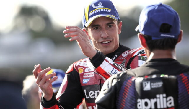 MotoGP rider in a red racing suit with a Michelin cap gestures while talking to a teammate in the pit area.