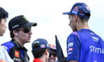 Two MotoGP riders in team gear chat outdoors, one wearing a black Yamaha cap with sunglasses and AirPods.