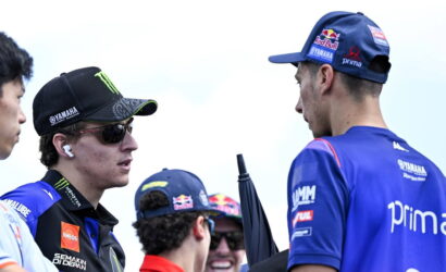 Two MotoGP riders in team gear chat outdoors, one wearing a black Yamaha cap with sunglasses and AirPods.