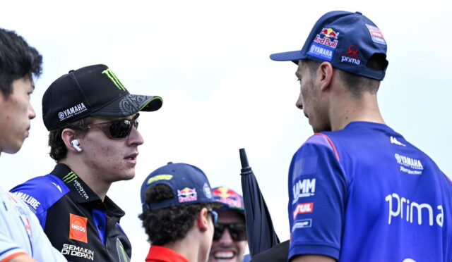 Two MotoGP riders in team gear chat outdoors, one wearing a black Yamaha cap with sunglasses and AirPods.