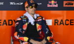 MotoGP rider in colorful KTM/Red Bull race suit sits in a red chair against an orange sponsor wall, wearing sunglasses and a cap with the Red Bull logo.