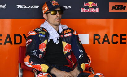 MotoGP rider in colorful KTM/Red Bull race suit sits in a red chair against an orange sponsor wall, wearing sunglasses and a cap with the Red Bull logo.