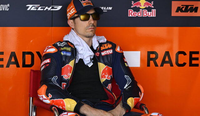 MotoGP rider in colorful KTM/Red Bull race suit sits in a red chair against an orange sponsor wall, wearing sunglasses and a cap with the Red Bull logo.