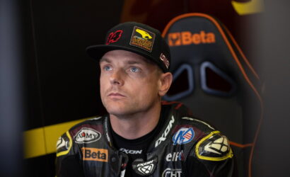 Male race car driver in a patched racing suit and cap, seated in a chair, gazing upward in thought.