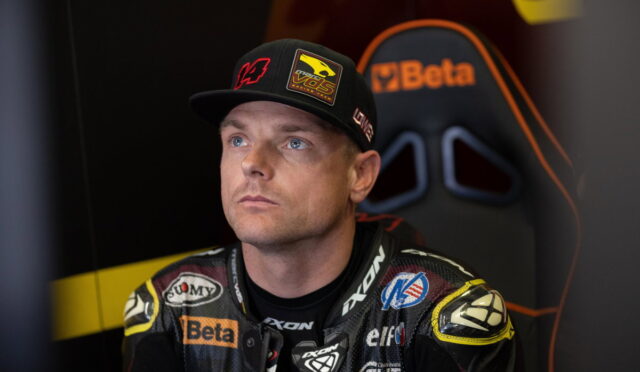 Male race car driver in a patched racing suit and cap, seated in a chair, gazing upward in thought.