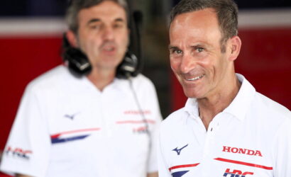 Smiling man in a white Honda polo shirt in the foreground with another team member wearing a headset in the background inside a red-paned pit area.