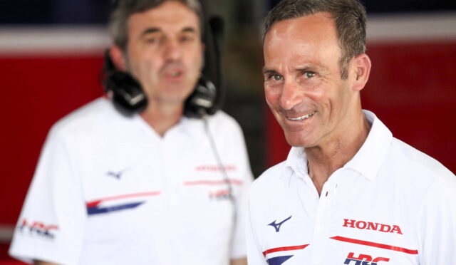 Smiling man in a white Honda polo shirt in the foreground with another team member wearing a headset in the background inside a red-paned pit area.