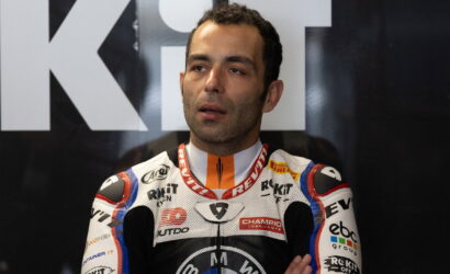 Moto racer in a sponsor-laden racing suit, standing with arms crossed, in front of a dark backdrop with large white letters behind him.