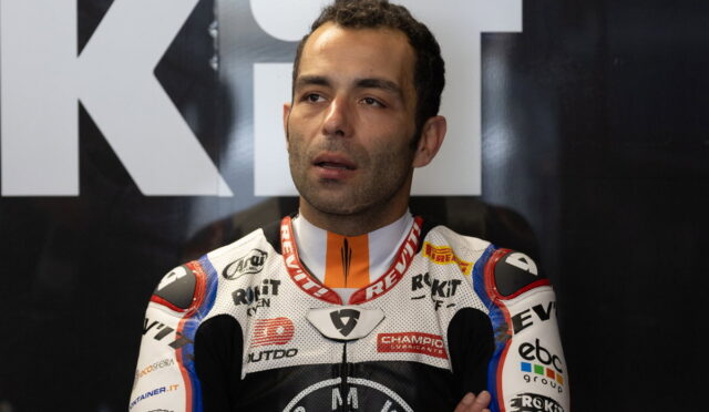 Moto racer in a sponsor-laden racing suit, standing with arms crossed, in front of a dark backdrop with large white letters behind him.