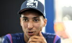 Close-up of a man wearing a navy cap with a '21.COM' logo, speaking with his hand near his chin.