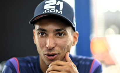 Close-up of a man wearing a navy cap with a '21.COM' logo, speaking with his hand near his chin.