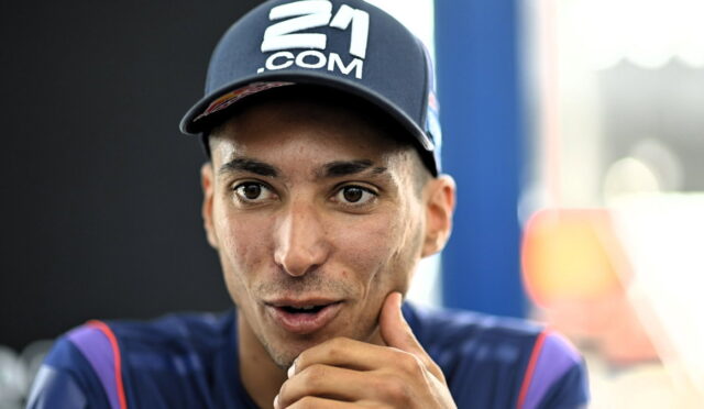 Close-up of a man wearing a navy cap with a '21.COM' logo, speaking with his hand near his chin.