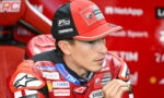 Close-up of a Ducati MotoGP rider in a red racing suit and cap with sponsor logos, seated in a red team area.