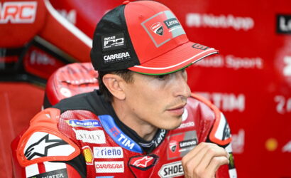 Close-up of a Ducati MotoGP rider in a red racing suit and cap with sponsor logos, seated in a red team area.