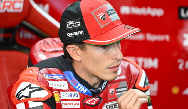 Close-up of a Ducati MotoGP rider in a red racing suit and cap with sponsor logos, seated in a red team area.