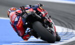 MotoGP rider leans into a sharp turn on a racetrack, red-blue suit and bike in motion beside blue-painted curbs.