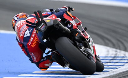 MotoGP rider leans into a sharp turn on a racetrack, red-blue suit and bike in motion beside blue-painted curbs.