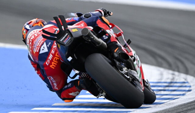 MotoGP rider leans into a sharp turn on a racetrack, red-blue suit and bike in motion beside blue-painted curbs.