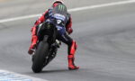 Moto racer in a red-and-blue suit leans into a corner on a Yamaha race bike with Monster logos on the suit and bike, on a track edge nearby safety lines.