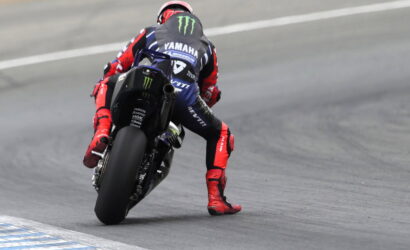 Moto racer in a red-and-blue suit leans into a corner on a Yamaha race bike with Monster logos on the suit and bike, on a track edge nearby safety lines.