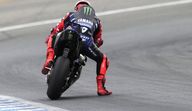 Moto racer in a red-and-blue suit leans into a corner on a Yamaha race bike with Monster logos on the suit and bike, on a track edge nearby safety lines.