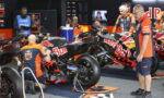 Mechanics in orange and blue Red Bull KTM uniforms service a KTM motorcycle in a pit garage.