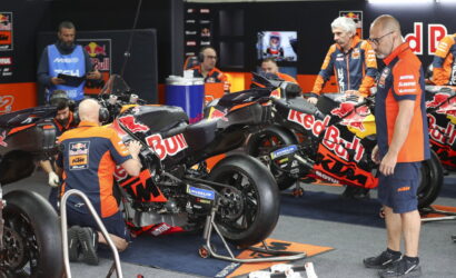 Mechanics in orange and blue Red Bull KTM uniforms service a KTM motorcycle in a pit garage.