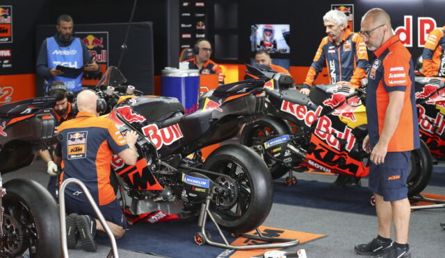 Mechanics in orange and blue Red Bull KTM uniforms service a KTM motorcycle in a pit garage.