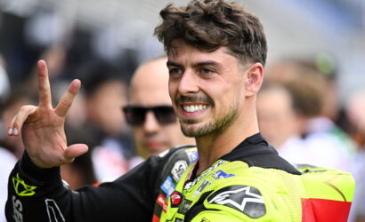 Male motorcyclist in neon-yellow racing suit smiling and flashing a peace sign at the camera, in a race paddock setting