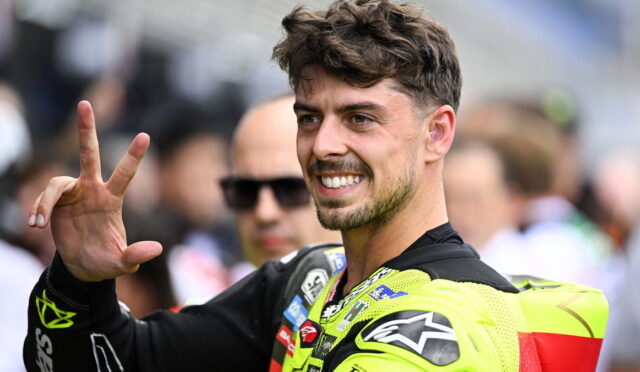 Male motorcyclist in neon-yellow racing suit smiling and flashing a peace sign at the camera, in a race paddock setting