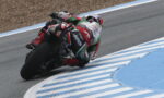 Motorcycle racer in a red, white, and green suit leaning into a sharp turn on a racetrack with blue and white curbs nearby (rear view).