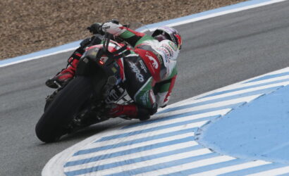 Motorcycle racer in a red, white, and green suit leaning into a sharp turn on a racetrack with blue and white curbs nearby (rear view).