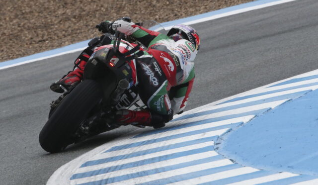 Motorcycle racer in a red, white, and green suit leaning into a sharp turn on a racetrack with blue and white curbs nearby (rear view).