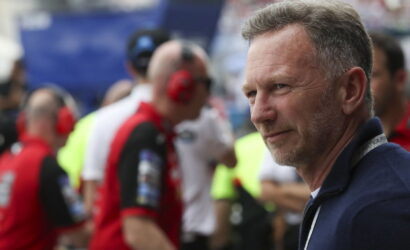 Man with graying hair in a navy jacket at a motorsport event, blurred pit crew in red in the background.