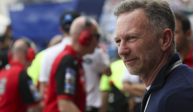 Man with graying hair in a navy jacket at a motorsport event, blurred pit crew in red in the background.