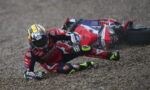 MotoGP rider in red leathers crashes on a gravel surface, helmeted, with a red sport bike on its side behind him.