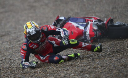 MotoGP rider in red leathers crashes on a gravel surface, helmeted, with a red sport bike on its side behind him.