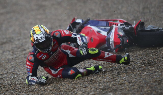 MotoGP rider in red leathers crashes on a gravel surface, helmeted, with a red sport bike on its side behind him.