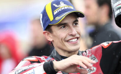 Person in a red racing suit and blue Michelin cap smiles while celebrating with arms raised.3c6️ Likely a motorsport winner or podium moment.