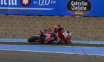 Two motorcycle racers in red gear crash on a blue-painted track, lying beside their bikes as they slide on the run-off area with a gravel barrier behind and sponsor banners above.