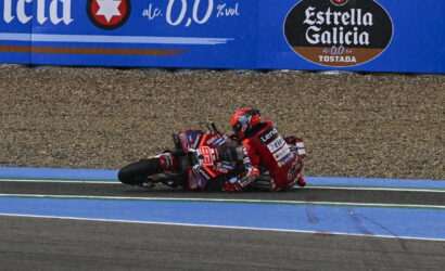 Two motorcycle racers in red gear crash on a blue-painted track, lying beside their bikes as they slide on the run-off area with a gravel barrier behind and sponsor banners above.