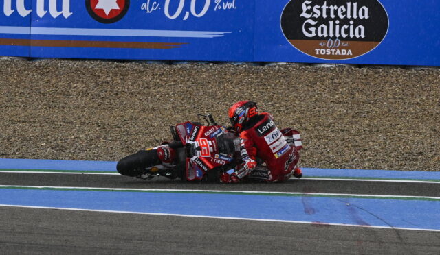 Two motorcycle racers in red gear crash on a blue-painted track, lying beside their bikes as they slide on the run-off area with a gravel barrier behind and sponsor banners above.