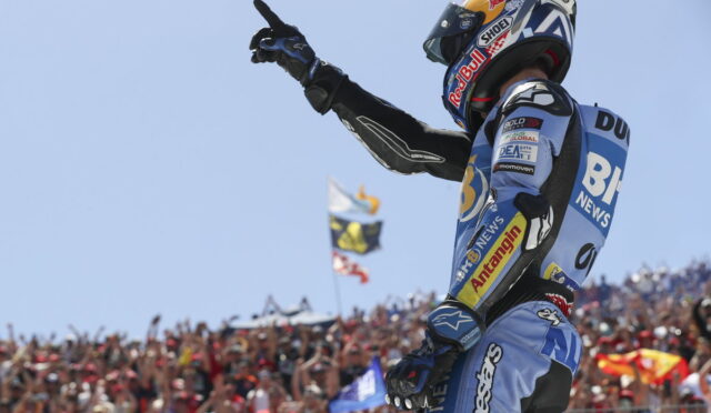 Motocross rider in blue racing suit and helmet points skyward as a cheering crowd waves flags in the background.