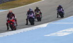 Four motorcycle racers in colorful gear ride side by side on a race track, with a blue run-off area on the right side.