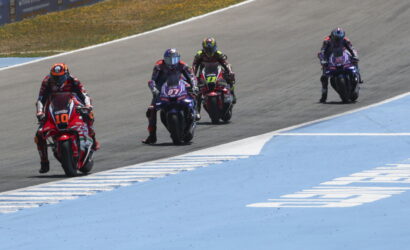 Four motorcycle racers in colorful gear ride side by side on a race track, with a blue run-off area on the right side.