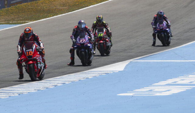 Four motorcycle racers in colorful gear ride side by side on a race track, with a blue run-off area on the right side.
