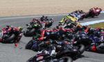 Group of racing motorcycles leaning into a tight corner on a track, riders in colorful gear close together.